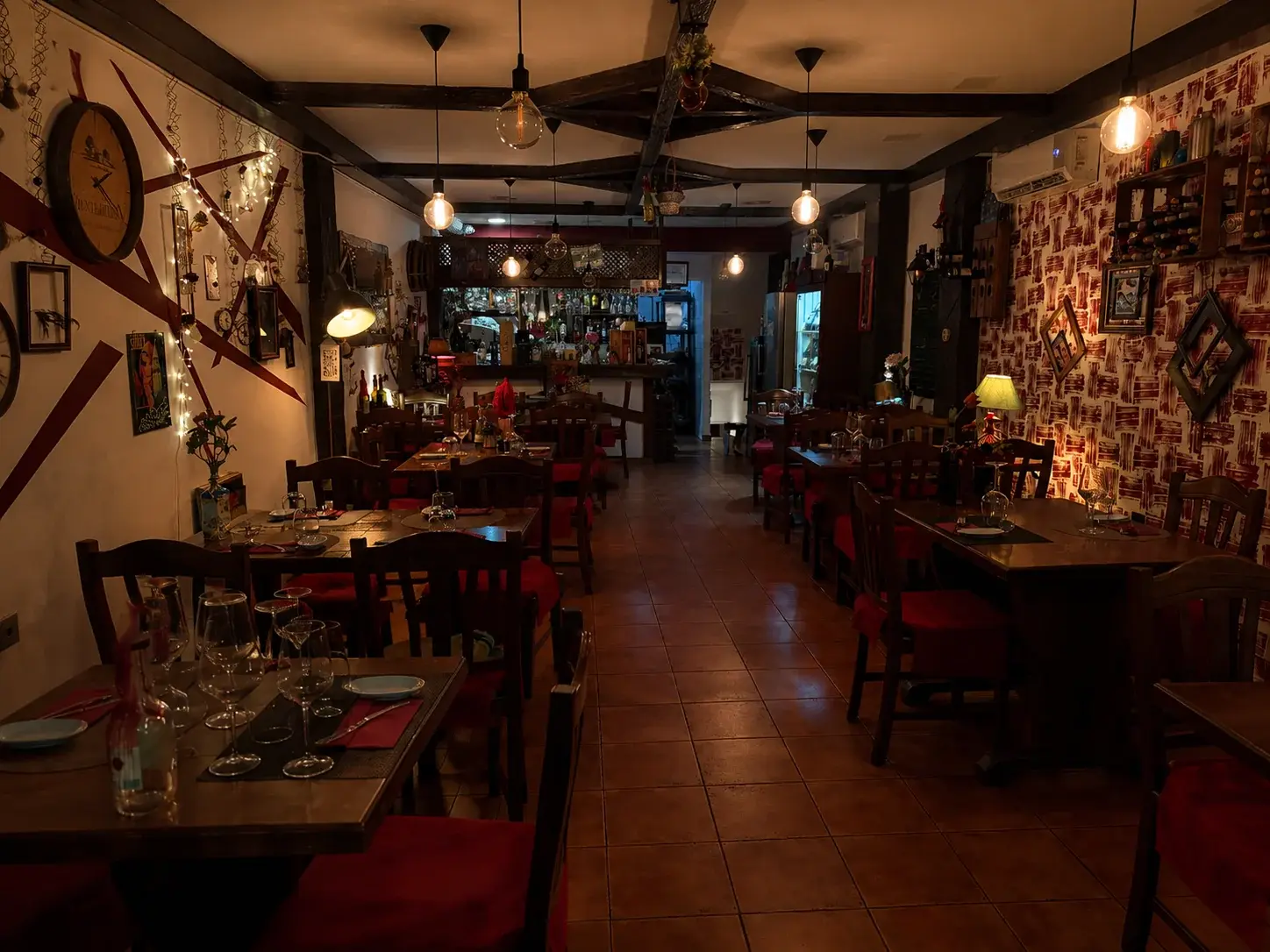 ARDI's long dining room in the evening: wooden tables and red-cushioned chairs under hanging bulb lights, with a tiled floor leading back to the bar.
